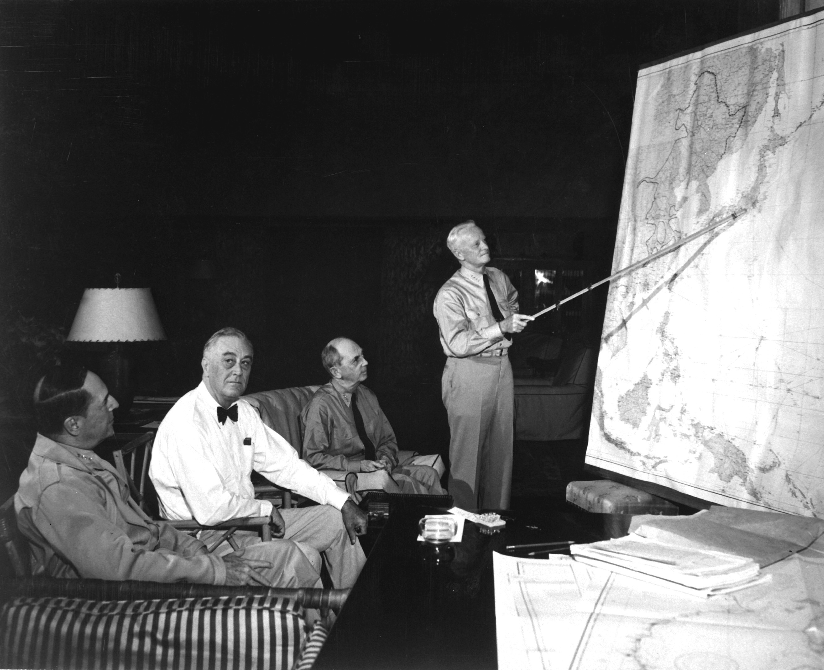 Conference in Hawaii, September 1944. Left to right: General MacArthur, President Roosevelt, Admiral Leahy, Admiral Nimitz.|alt=Three men are seated in lounge chairs. One is standing, holding a long stick and pointing to the location of Japan on a wall map of the Pacific.