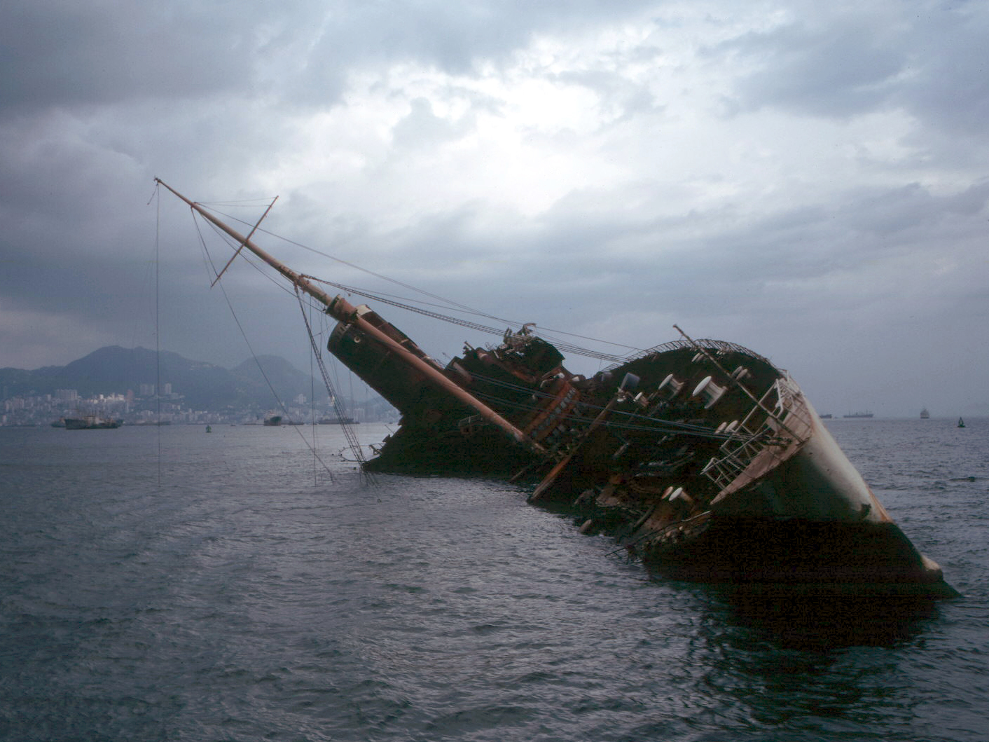 1972: The wreck of the Seawise University, the former RMS Queen Elizabeth, in Victoria Harbour, Hong Kong.|alt= A half-sunken ship lies listing at 45 degrees