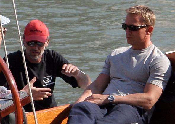 Craig in Venice during filming.|alt=A man wearing a grey shirt, jeans and sunglasses sits on a boat at sea. Besides him, a man wearing a black shirt, red cap and sunglasses talks to another which is mostly off the picture.