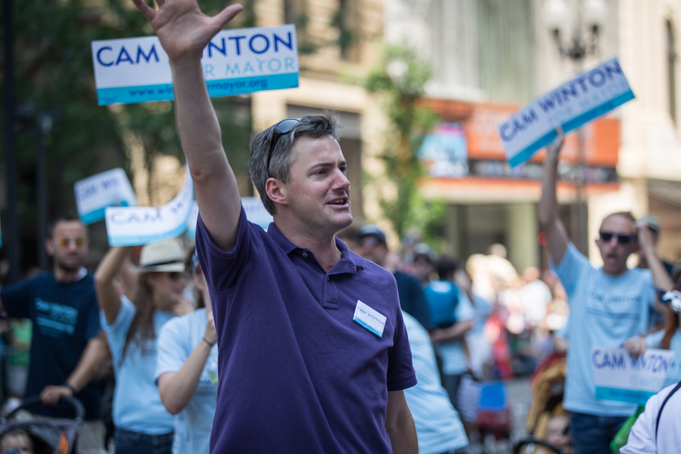 Winton campaigning for mayor at the 2013 Twin Cities Pride Parade |alt=A man in his mid-30s wearing a purple polo shirt waves as people behind him hold up signs with his name, Cam Winton.